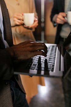 Close-up of hands typing on a laptop with colleagues holding coffee cups.