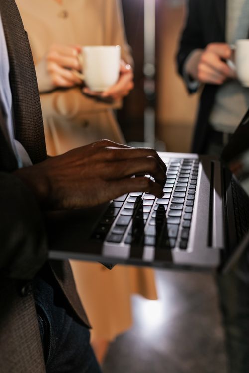 Free Close-up of hands typing on a laptop with colleagues holding coffee cups. Stock Photo
