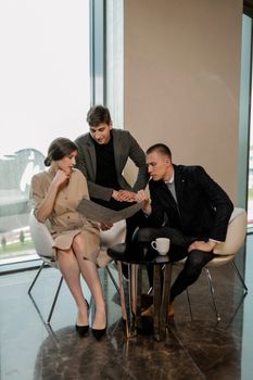 Three colleagues having a focused discussion in an office setting by a large window.