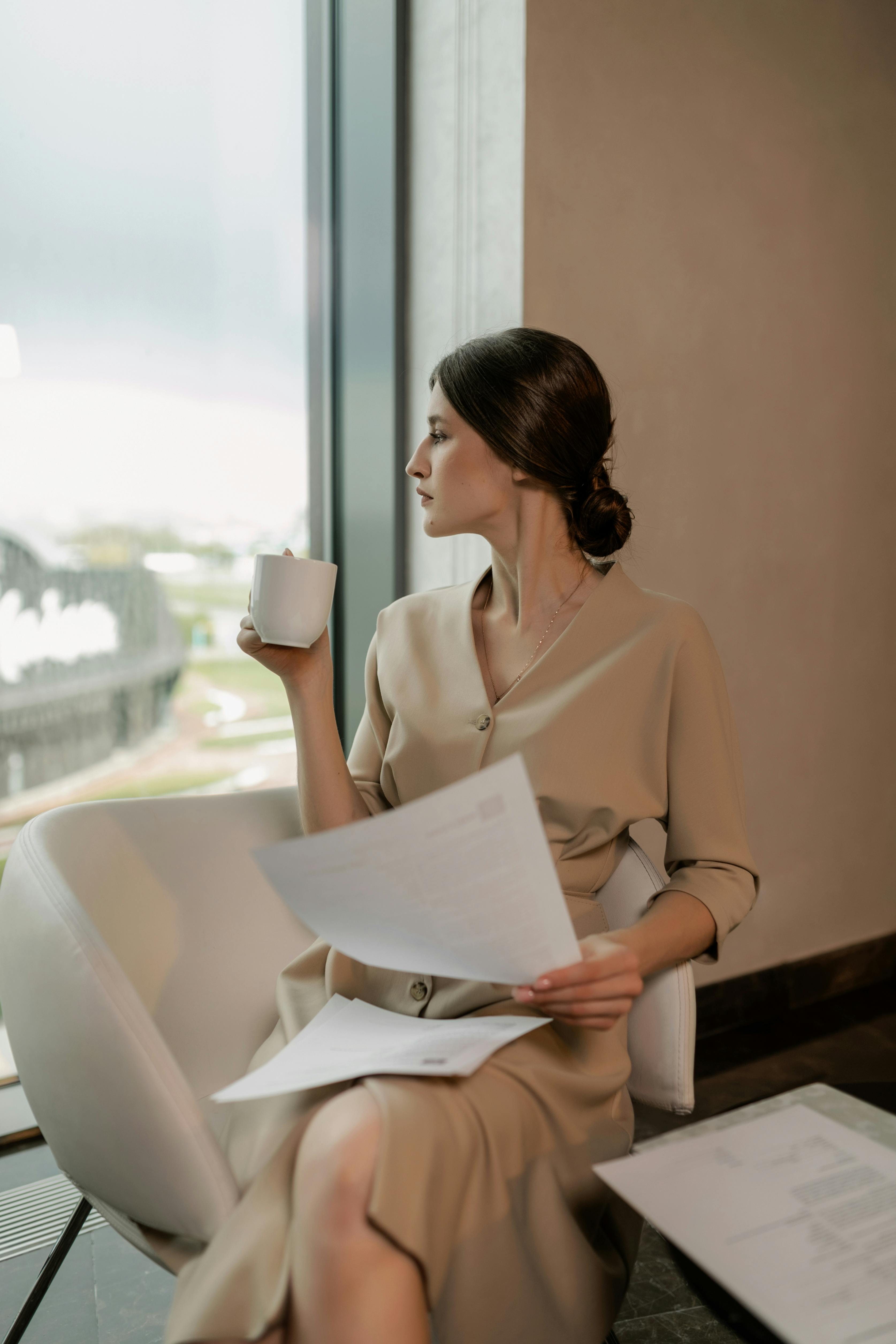 a woman in beige dress sitting on the chair while holding a cup of coffee