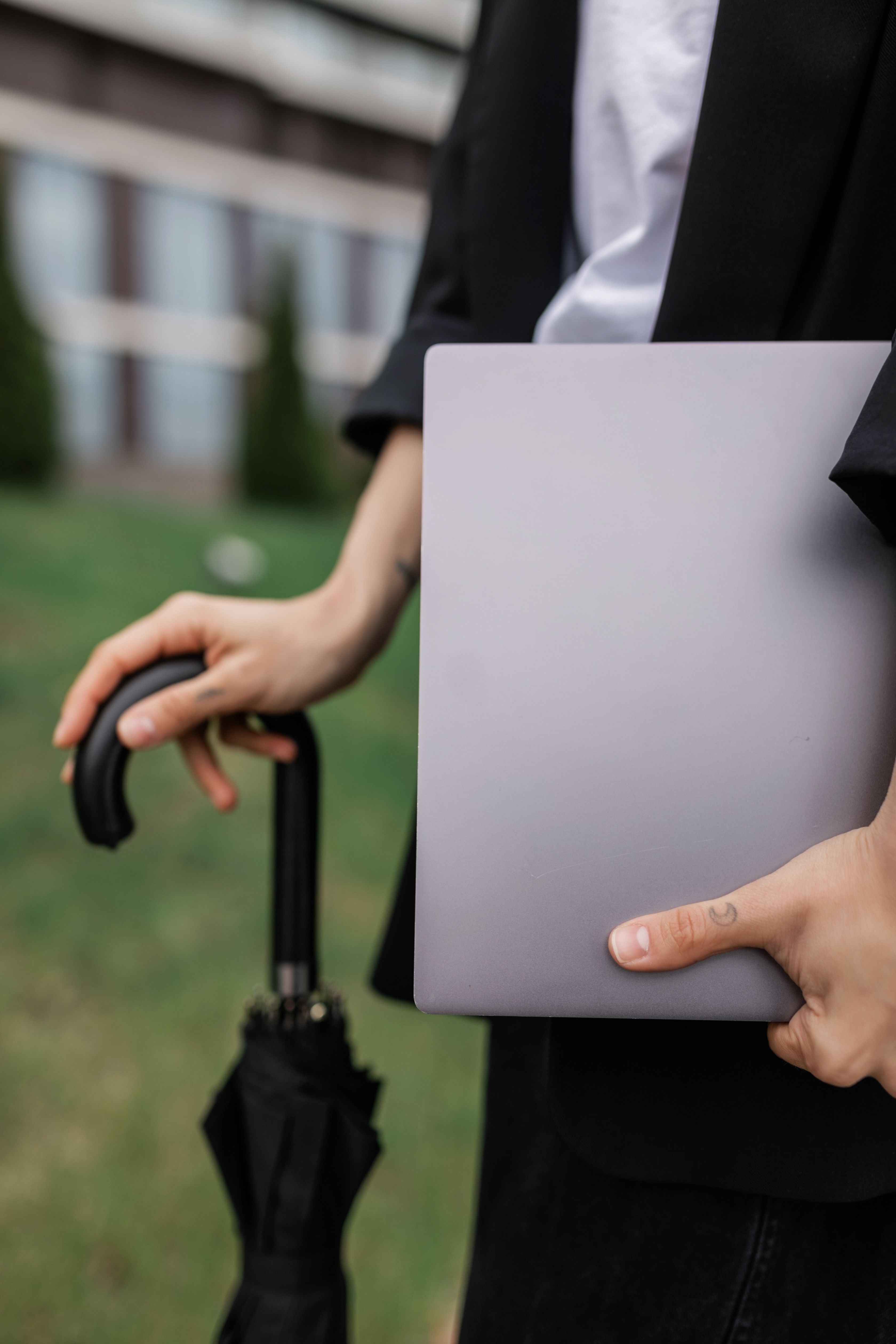 A person in business attire holding a laptop and umbrella, captured outdoors.