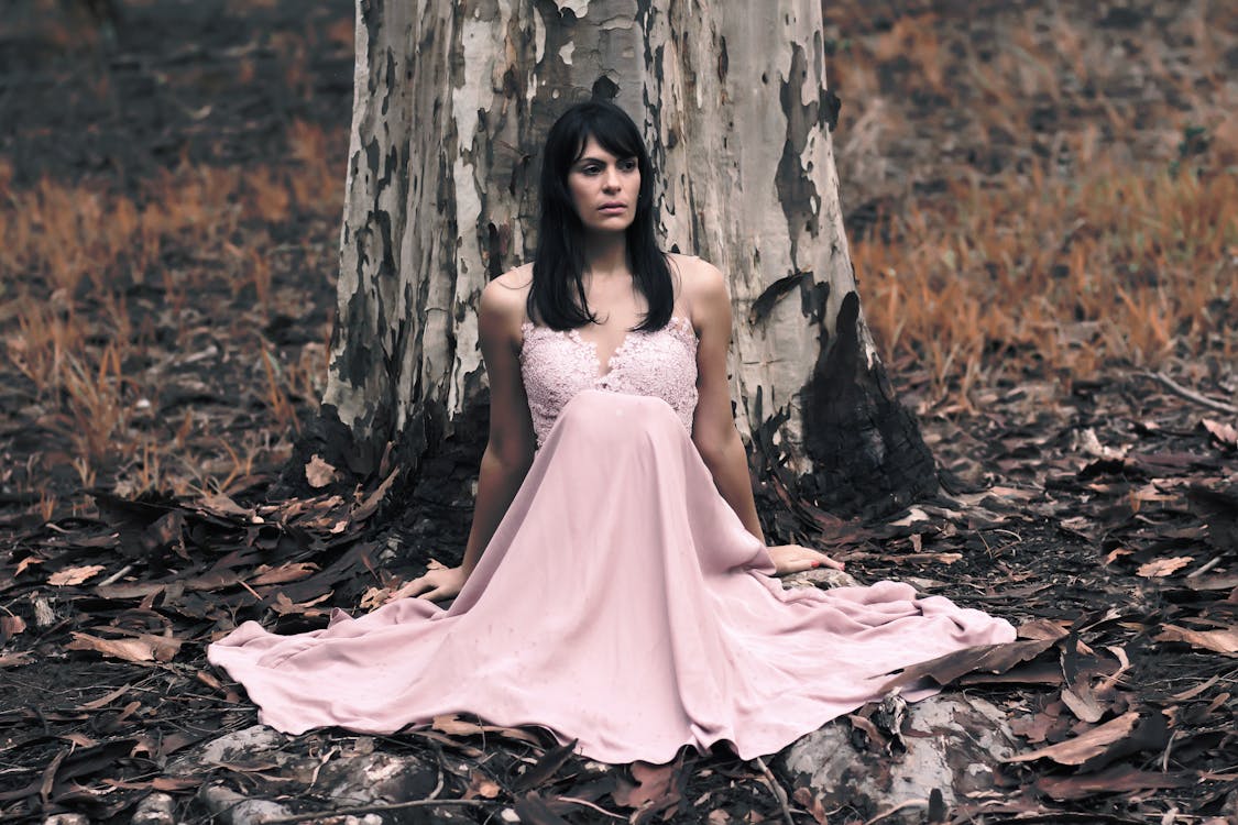 나무 앞에 앉아 분홍색 민소매 드레스에 검은 머리 여자의 얕은 초점 사진