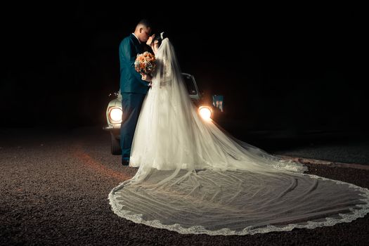 A couple shares an intimate moment on their wedding night, illuminated by a vintage car's headlights.