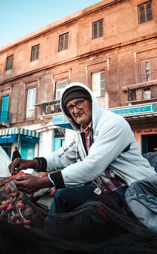 Free Elderly man in casual attire mending a fishing net outdoors in front of a historic building. Stock Photo
