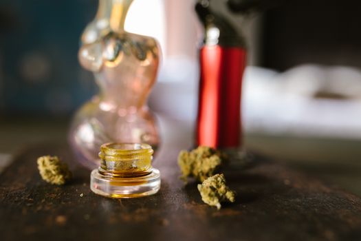 Glass jar with cannabis concentrate, surrounded by dried marijuana buds and smoking accessories on a table.