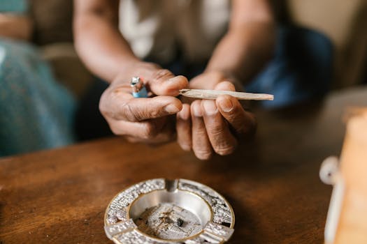Close-up of hands holding a cannabis joint with an ashtray on a wooden table.