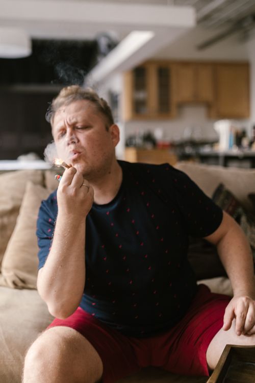 Free Man seated on a sofa in a living room, casually smoking, representing a moment of relaxation. Stock Photo