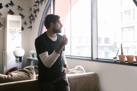 Man indoors by a window holding a vape, surrounded by decor and soft lighting.