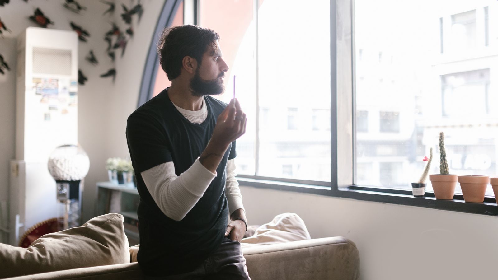 Man indoors by a window holding a vape, surrounded by decor and soft lighting.