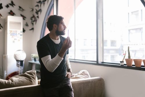 Free Man indoors by a window holding a vape, surrounded by decor and soft lighting. Stock Photo