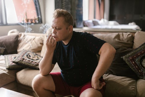 Free Man in Black Crew Neck T-shirt and Red Shorts Sitting on Couch and Smoking Stock Photo