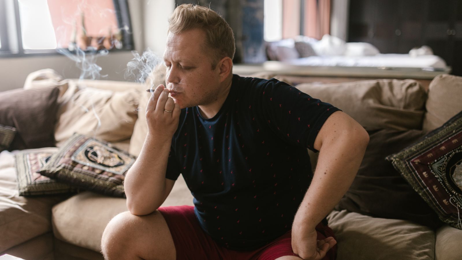 Adult man sitting indoors, smoking a joint, in a relaxed setting.