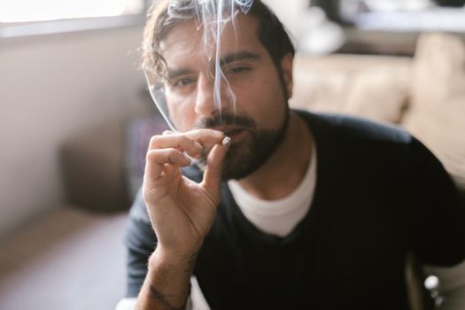 Man smoking a cigarette indoors with a blurry background, emitting visible smoke.
