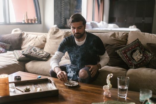 Young man enjoying aromatic smoke in a cozy living room setting with incense and a pipe.