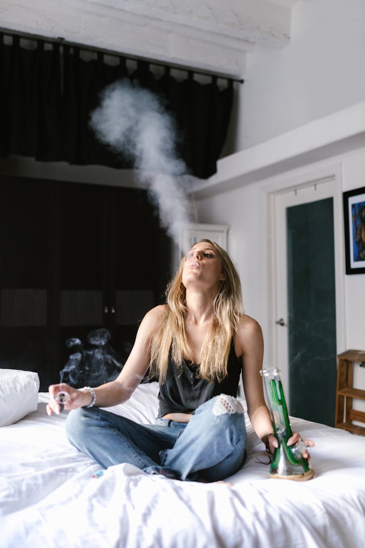 A Woman In Black Tank Top And Denim Jeans Sitting On The Bed While Smoking Weed