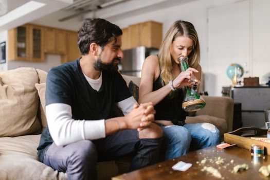 Two adults enjoying a leisure moment in a cozy home setting, one using a bong with cannabis.