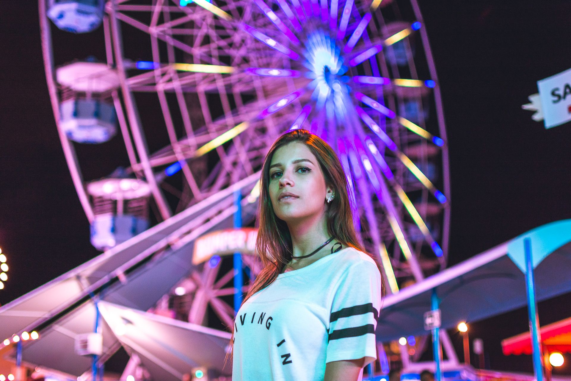 Portrait of a young woman standing at a vibrant nighttime carnival with a Ferris wheel.