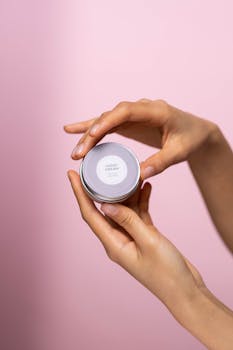 Close-up of elegant hands holding a jar of hand cream in a soft pink background, emphasizing skincare and beauty.