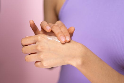 Close-up of a woman applying moisturizer to her hand against a pink background.
