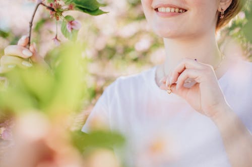 Free Crop unrecognizable female with golden pendant standing near blooming branches of tree with green leaves in garden on blurred background Stock Photo