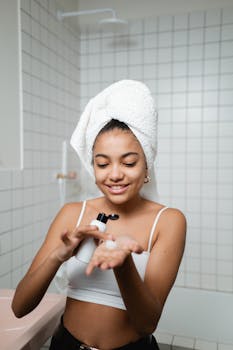 Young woman with towel on head applies lotion as part of her daily skincare routine in a contemporary bathroom.