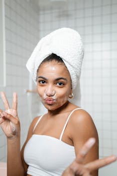 A young woman applies skincare while giving a peace sign, showcasing her morning beauty routine in a bathroom.