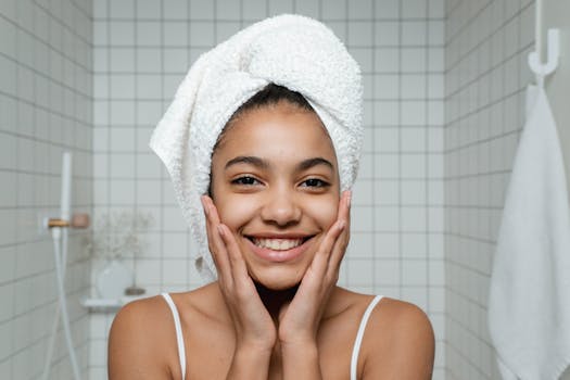 A young woman smiles during her morning skincare routine with a towel around her head.