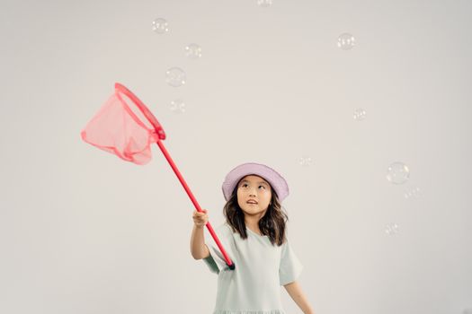 A young girl with a butterfly net catching bubbles against a white background, wearing a bucket hat.