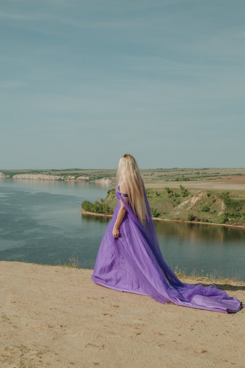 Back view of unrecognizable elegant woman with long blond hair in violet dress contemplating river from shore under cloudy sky