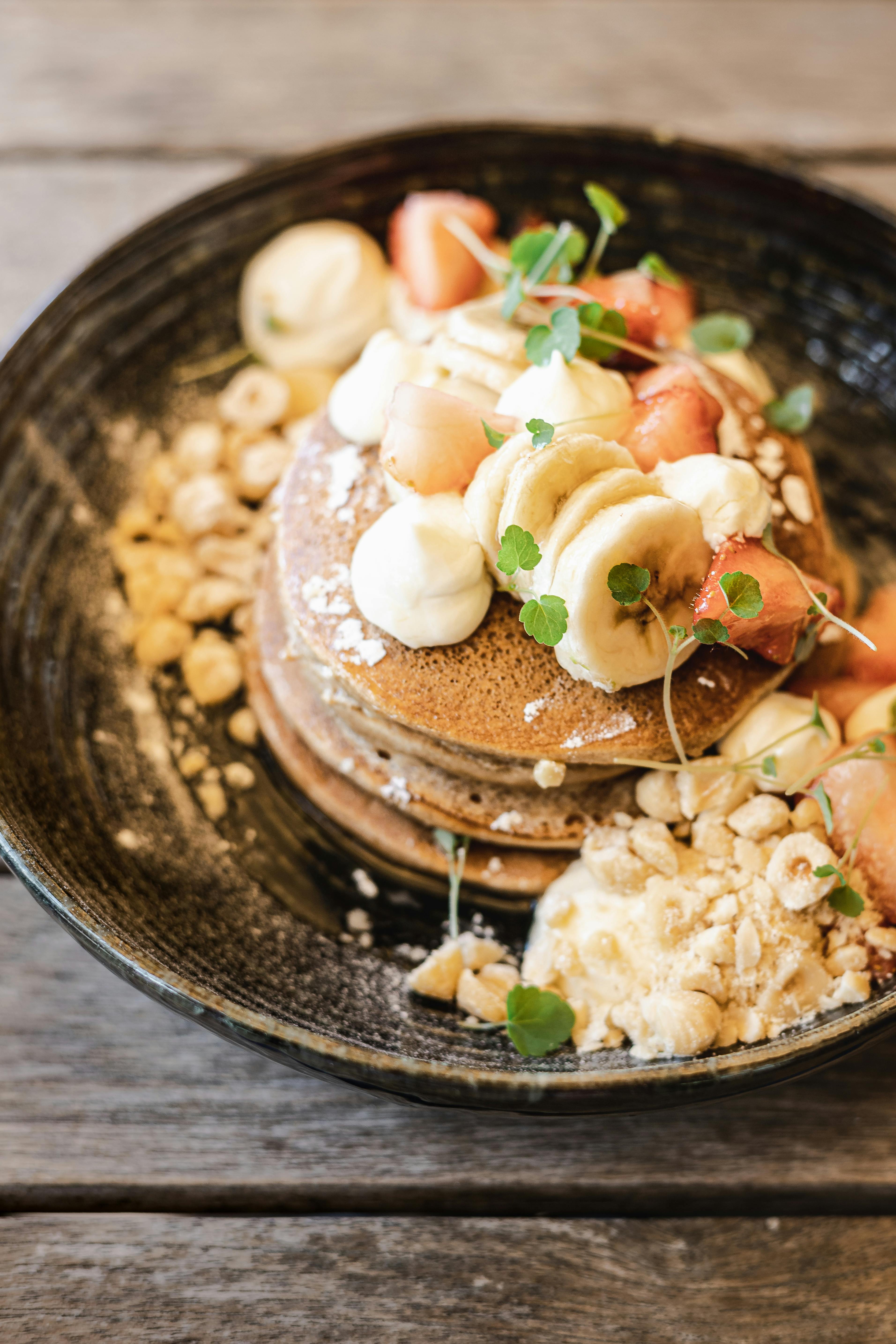 Stack of pancakes topped with bananas, strawberries, cream, and nuts on a rustic plate.