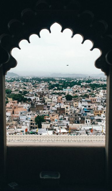 Beautiful cityscape of Udaipur, India seen through a traditional arched window, showcasing the vibrant city and cloudy sky.