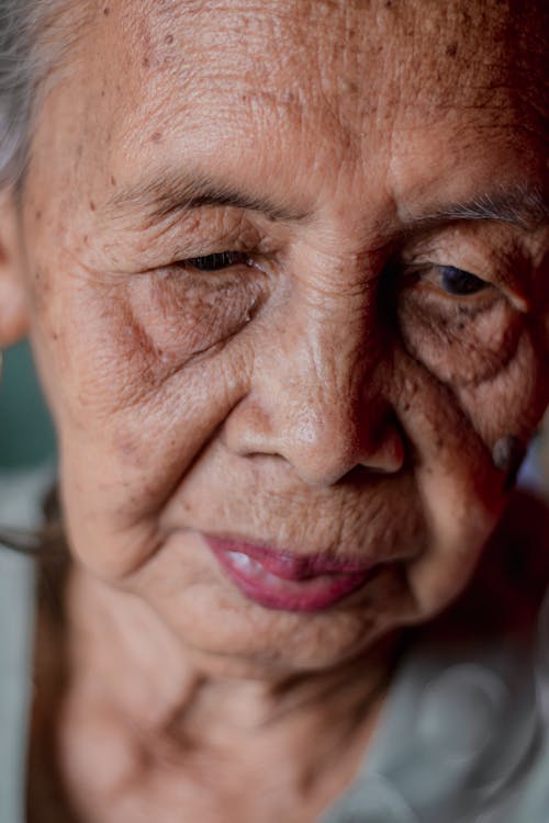 Crop elderly woman with wrinkles and gray hair looking down against blurred background in daytime