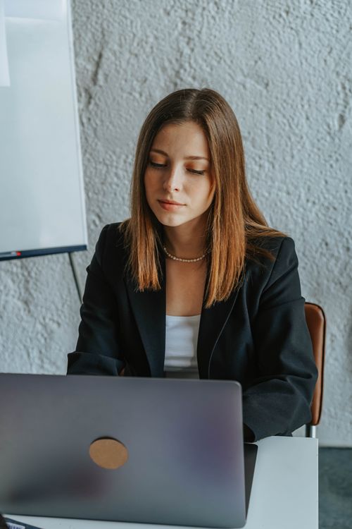 Free Woman in a black blazer working at a laptop in an indoor office setting. Stock Photo