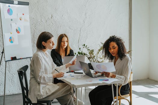 Three women collaborating at a workplace with laptops and charts, highlighting teamwork and diversity.