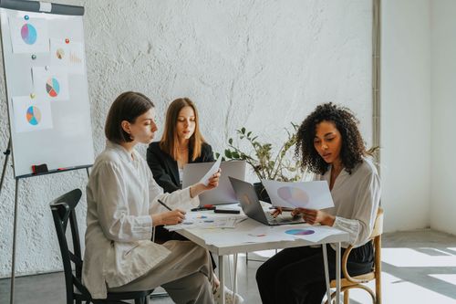 Free Three women collaborating at a workplace with laptops and charts, highlighting teamwork and diversity. Stock Photo