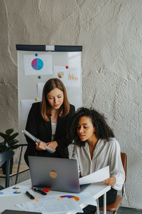 Free Two professional women discussing work at an office desk with a laptop and paperwork. Stock Photo