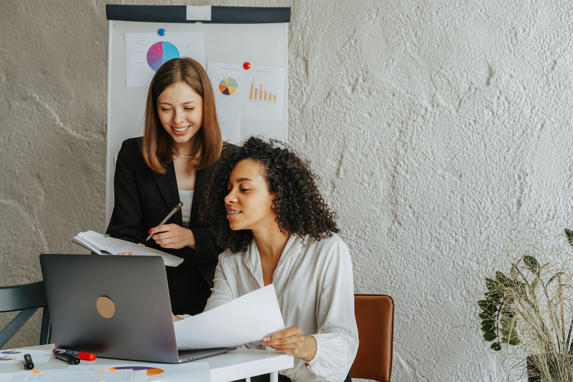 Two women discussing business strategy at the office using a laptop and charts.