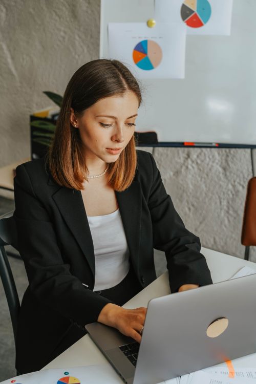 Free Confident businesswoman working on a laptop in a modern office environment with charts in the background. Stock Photo