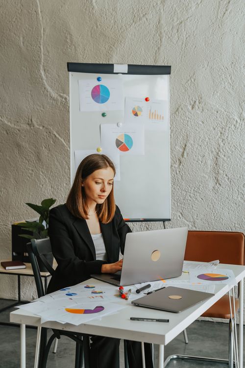 Free Professional woman in blazer analyzing charts and graphs on laptop in modern office setting. Stock Photo
