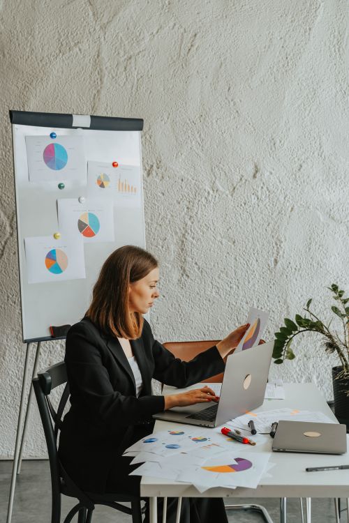 Free A professional woman examines charts at her desk, working on a laptop and reviewing papers. Stock Photo