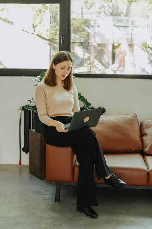 Free Businesswoman sitting on a sofa, working on laptop in a sunny office. Ideal for business and remote work themes. Stock Photo