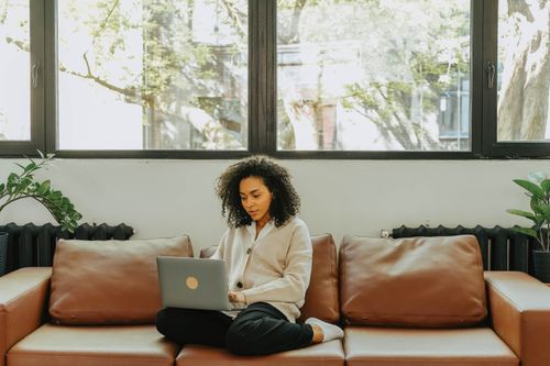 Free Woman with curly hair working on a laptop while sitting on a brown sofa in a bright living room. Stock Photo