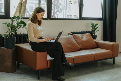 Free A professional woman working on a laptop, seated on a comfortable couch with indoor plants. Stock Photo