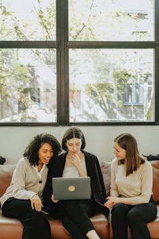 Three diverse businesswomen sitting together on a couch, collaborating on a laptop in a bright office setting.
