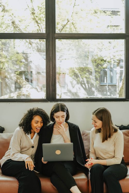Free Three diverse businesswomen sitting together on a couch, collaborating on a laptop in a bright office setting. Stock Photo