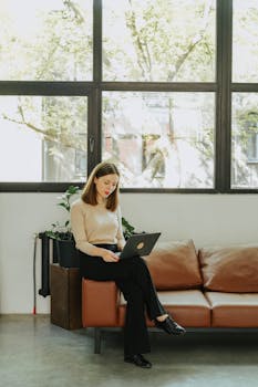 A woman working on a laptop while sitting on a sofa in a bright, modern living room with large windows.