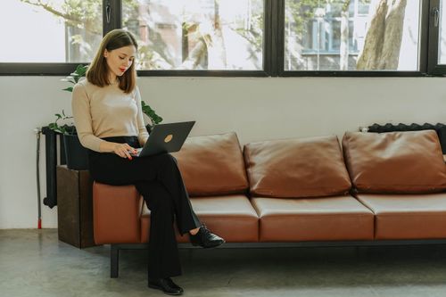Free A young woman working on a laptop while sitting on a comfortable sofa in a well-lit modern room. Stock Photo
