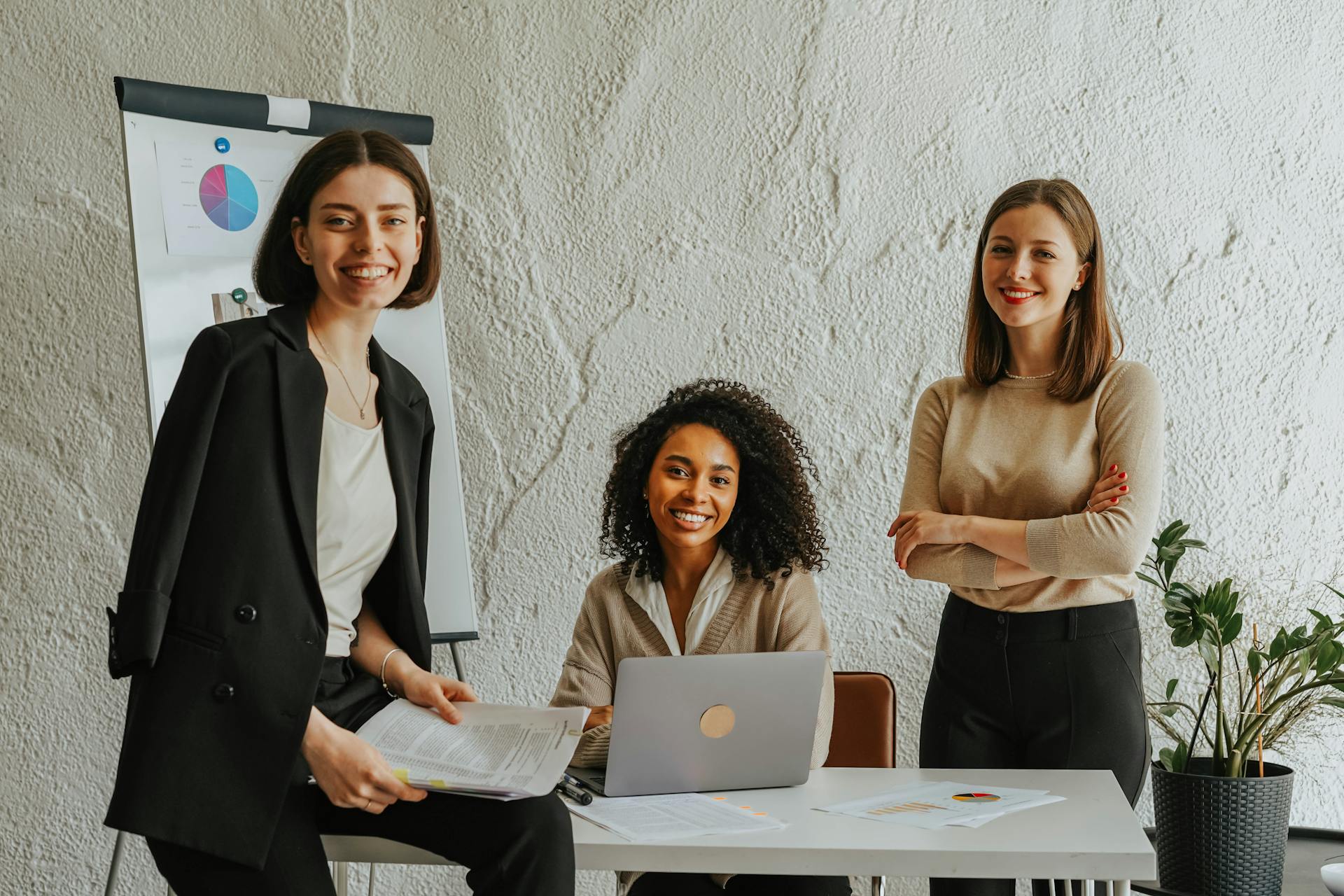 Three businesswomen meeting at a table, using a laptop and presenting charts in a modern office.