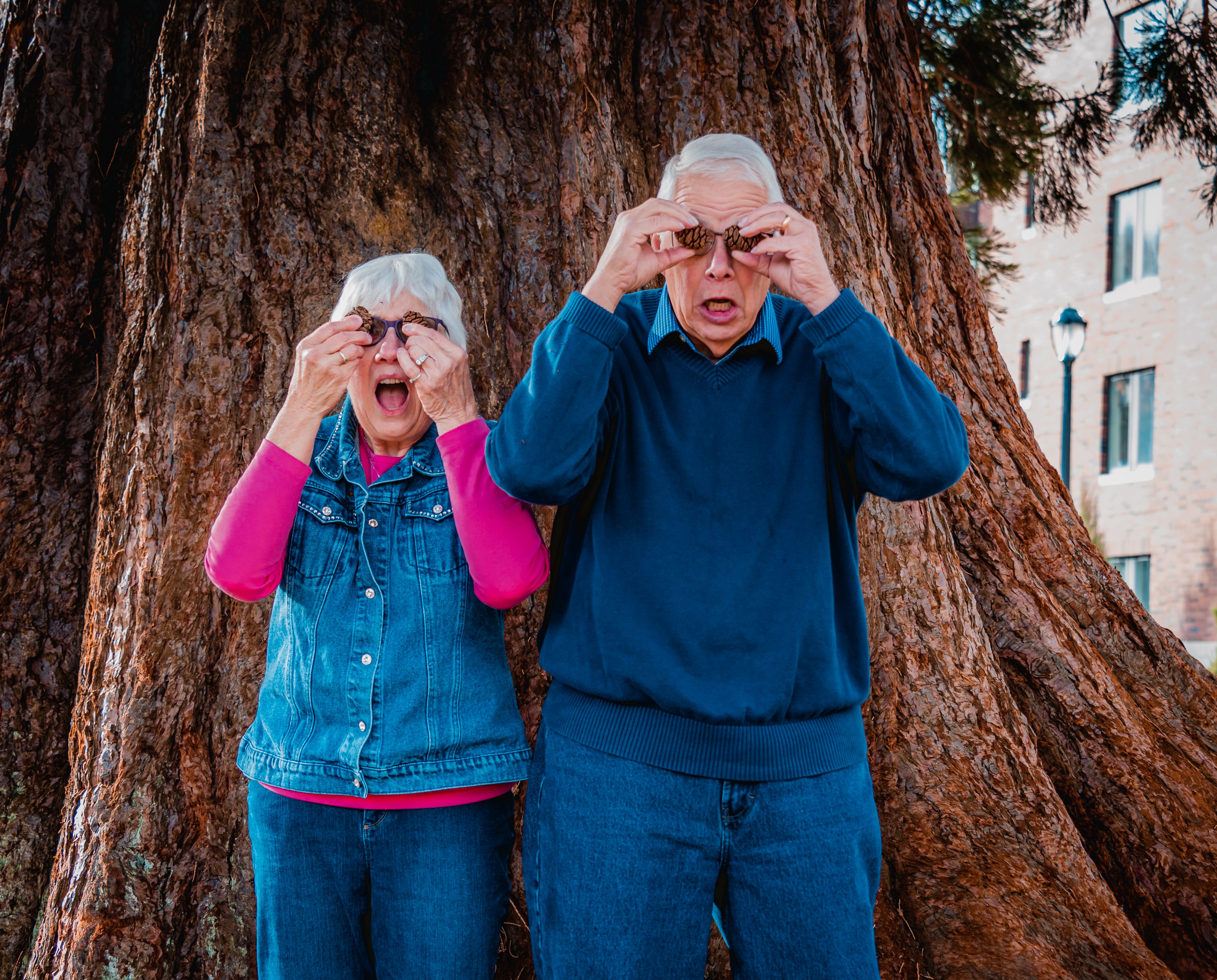 An elderly couple playfully using pinecones as glasses in front of a large tree trunk.