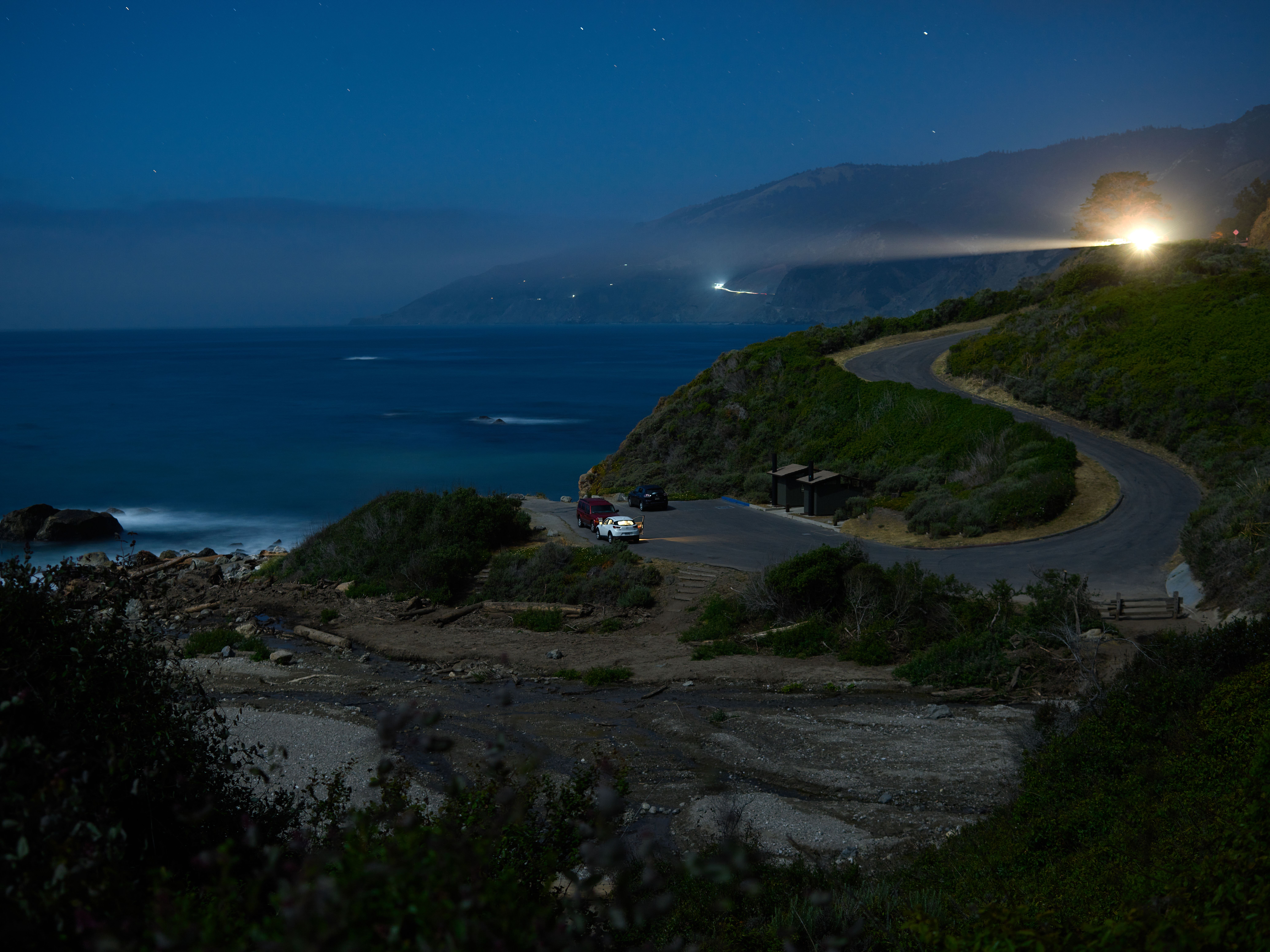 Dramatic night view of a coastal road under a star-filled sky, featuring ocean and light trails.